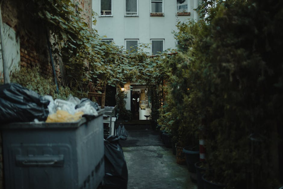 A narrow outdoor alleyway between residential buildings, featuring a large grey plastic dumpster overflowing with black and white rubbish bags on the left side, with some bags sitting atop the open lid. The ground surface is a worn, dark paved area with visible patches of dirt and debris. On the right side, a row of large potted plants and shrubs extends along the length of the alley, with dense greenery and foliage partially obscuring the view. At the end of the alley, there is a small, illuminated entrance with a door, framed by alley walls covered in climbing vines and creeping plant foliage. Overhead, a tangle of vines and branches creates a canopy, with some leaves spilling into the scene. In the background, a multi-storey white residential building with several windows is visible, providing a stark contrast to the natural greenery and the utilitarian rubbish collection area. The overall scene depicts an urban outdoor back alley used for waste disposal, highlighting the importance of professional rubbish removal services like those offered by Waste Removal Brixton to maintain cleanliness and proper waste management in residential areas.
