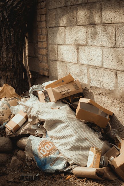 A pile of mixed waste materials is seen against a rough, unfinished concrete and brick wall, situated outdoors near a tree trunk. The collection includes flattened cardboard boxes with printed labels, some partially torn or crumpled, alongside discarded paper and plastic packaging. A large beige and white sack with a textured surface holds some of the waste, resting on uneven ground composed of small rocks and dirt. The cardboard boxes vary in size, with some open and others closed, revealing no contents. Shadows cast by the nearby tree suggest natural daylight, and the scene typifies privately collected rubbish, likely managed by a waste removal service such as Waste Removal Brixton, involving on-site waste clearance in a residential or back-alley environment.