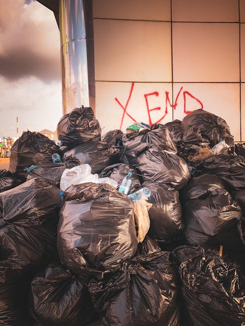 A large pile of black rubbish bags, some torn or partially open, stacked against a modern building with reflective panels on a street or public area. Several discarded plastic water bottles are visible among the bags, which are placed close to a graffiti-covered wall featuring red spray paint spelling 'XEND'. The environment appears to be an outdoor urban space during daylight, with adjacent buildings and a cloudy sky visible in the background. The scene suggests an instance of waste accumulation awaiting collection, exemplifying private or independent rubbish disposal that waste removal services like Waste Removal Brixton might handle, especially when managing excess or irregular waste in a commercial or residential setting. The textured black plastic bags indicate typical refuse containment, and their placement indicates an active effort of waste disposal or clearance in the area.