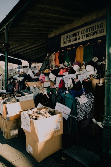 The image shows an outdoor market stall with a variety of second-hand clothing and accessories arranged on tables, shelves, and hanging displays beneath a dark, corrugated roof structure. The clothing includes sweaters, jackets, and hats in assorted colors such as black, white, pink, green, and orange, with some items positioned on mannequins or hung on racks. Cardboard boxes filled with additional items are placed on the ground, with labels displaying prices like £2.99 and £3.99. The stall bears a sign reading 'DISCOUNT CLOTHING FOOTWEAR & UNDERWEAR,' indicating that it primarily sells used apparel and footwear. Surrounding the stall is a paved area, likely part of an outdoor market or street fair, where waste removal services from Brixton are sometimes employed for clearing unsold or discarded goods. The lighting suggests natural daylight, casting shadows and illuminating the colorful fabric items. Overall, the scene captures a typical secondary market setup with an emphasis on pre-owned clothing sale and the potential for private collection or waste handling related to retail or market activity.