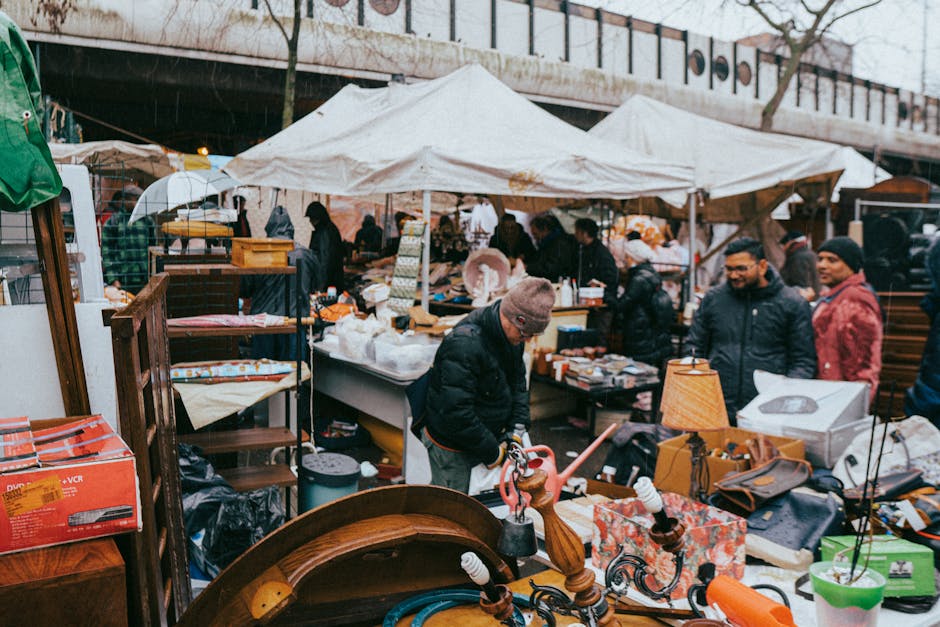 The image shows an outdoor market stall with a variety of second-hand clothing and accessories arranged on tables, shelves, and hanging displays beneath a dark, corrugated roof structure. The clothing includes sweaters, jackets, and hats in assorted colors such as black, white, pink, green, and orange, with some items positioned on mannequins or hung on racks. Cardboard boxes filled with additional items are placed on the ground, with labels displaying prices like £2.99 and £3.99. The stall bears a sign reading 'DISCOUNT CLOTHING FOOTWEAR & UNDERWEAR,' indicating that it primarily sells used apparel and footwear. Surrounding the stall is a paved area, likely part of an outdoor market or street fair, where waste removal services from Brixton are sometimes employed for clearing unsold or discarded goods. The lighting suggests natural daylight, casting shadows and illuminating the colorful fabric items. Overall, the scene captures a typical secondary market setup with an emphasis on pre-owned clothing sale and the potential for private collection or waste handling related to retail or market activity.