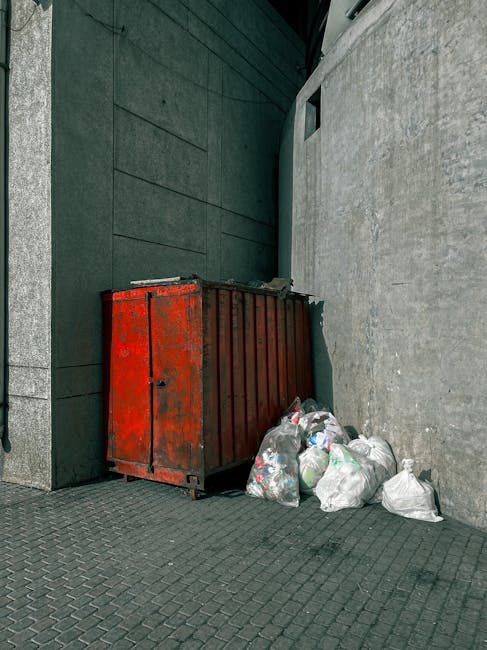 A large, red metal skip with visible rust and chipped paint is positioned against the junction of two building walls on a paved pavement. The skip is rectangular with vertical ridges on its sides and a flat lid on top. Surrounding the base of the skip are several tightly packed black and white plastic rubbish bags, some appearing slightly squashed or overfilled, containing various waste materials. One of the bags has a green plastic bottle visible inside. The background features a dark grey, smooth wall with horizontal paneling on the left, and a lighter grey, rough-textured wall on the right, which extends upwards and partially out of view. The pavement consists of small, interlocking stone tiles, and a narrow alleyway or corner is visible, suggesting an urban environment suitable for private waste collection and rubbish disposal services, such as those provided by Waste Removal Brixton.