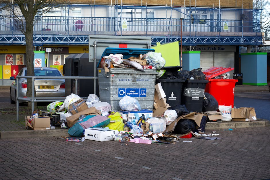 An outdoor scene showing a large accumulation of mixed waste materials, including cardboard boxes, paper bags, plastic bags, and loose packaging, overflowing from several large commercial rubbish bins positioned on a paved area near a roadside. The waste spills onto the surrounding pavement, with some items scattered on the ground. The bins are of various colours: a grey bin designated for mixed paper and cards, black bins for general waste, and a bright red bin, all placed against a metal fencing barrier. Behind the bins, there are commercial buildings with storefronts and signage, as well as a parked silver car to the left. The background features a building under construction or renovation with scaffolding covered in blue safety netting, and a leafless tree to the left. Natural daylight illuminates the scene, highlighting the disorganized state typical of bin overflow, illustrating an example of waste that may require professional rubbish removal services such as those offered by Waste Removal Brixton.