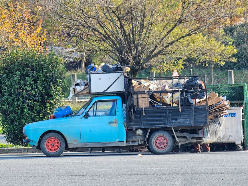 A small, older model pickup truck painted in faded blue with red wheels is parked alongside a road, loaded with a large amount of mixed household rubbish. The load includes flattened cardboard boxes, plastic bags filled with unknown items, and various debris stacked haphazardly, secured with ropes and tied-down materials. The truck's bed features metal side panels, some bent or showing signs of wear, and the cargo extends beyond the vehicle's rear tailgate. Behind the truck, a lush green hedge and a large tree with sparse yellow-green leaves indicate an outdoor environment, likely in a suburban or urban area during autumn. The pavement and curb are visible in the foreground, with a faint shadow cast by the truck suggesting daylight. This scene is indicative of private waste clearance or alternative rubbish removal, similar to services provided by Waste Removal Brixton, emphasizing the collection of bulky waste items outside of standard municipal collections.