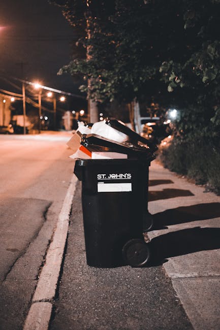 A black wheeled rubbish bin positioned on a sidewalk near the edge of a street at night. The bin has the label 'ST. JOHN'S' and is filled with various waste items, including cardboard, paper, and plastic. The cardboard appears slightly crumpled and is protruding from the top of the bin, which is situated next to a curb and bordered by a concrete pavement. Behind the bin, there are streetlights illuminating the roadway, and a row of trees and hedges line the sidewalk on the right side. In the background, parked cars are visible along the street, with some distant artificial lighting and power lines overhead. The scene suggests an urban environment where private waste collection services, such as waste removal Brixton, might handle rubbish disposal efficiently outside official local authority provisions, especially during evening hours when street lighting creates a warm, subdued atmosphere.